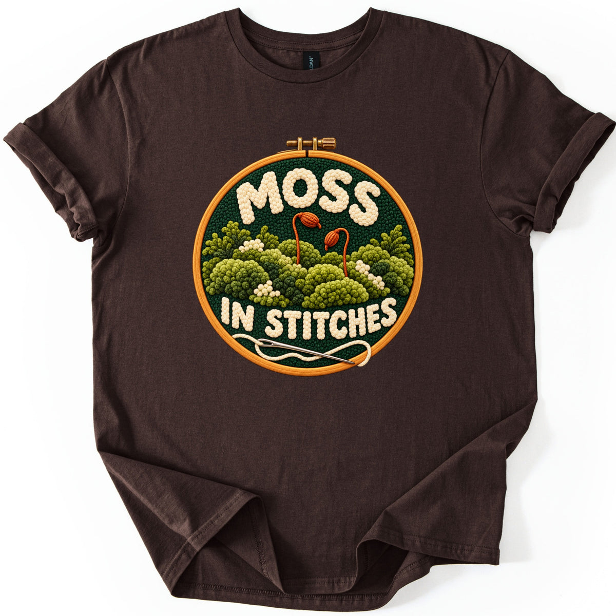 Moss in Stitches Comfort Colors T-shirt in Dark Chocolate, laid flat. A stitched embroidery hoop design of green moss clumps with two orange spore capsules, the words MOSS IN STITCHES in cream yarn, and a needle trailing thread. Sleeves rolled, on a white background.