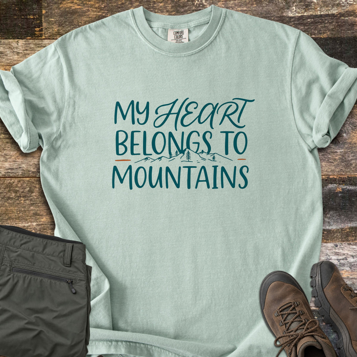 My Heart Belongs to Mountains Comfort Colors T-shirt in Bay, laid flat. Dark teal lettering reads My Heart Belongs to Mountains, with Heart in script, a line drawing of peaks and pines, and small rust-orange dashes. Olive pants and brown hiking boots sit on weathered wood.