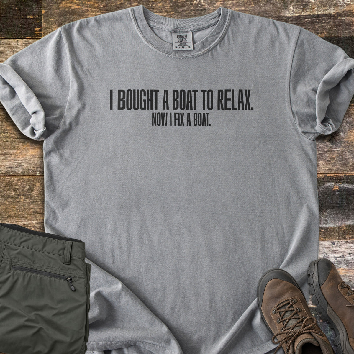 Boat To Relax Comfort Colors T-shirt in Grey, laid flat. Black block letters read I BOUGHT A BOAT TO RELAX. with NOW I FIX A BOAT. in smaller type below. Sleeves are cuffed, with folded olive cargo pants and brown hiking boots on weathered wood planks.
