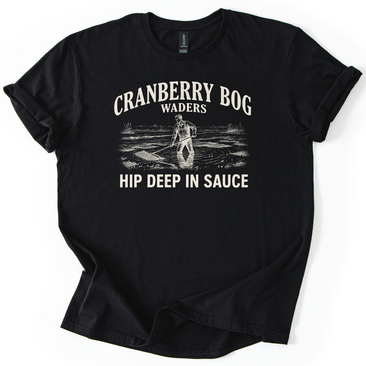 Cranberry Bog Waders Comfort Colors T-shirt in Black, laid flat. A cream etched drawing of a man in overalls standing hip deep in a flooded bog, raking floating cranberries, under the words Cranberry Bog Waders and above Hip Deep in Sauce. Sleeves cuffed, on white.
