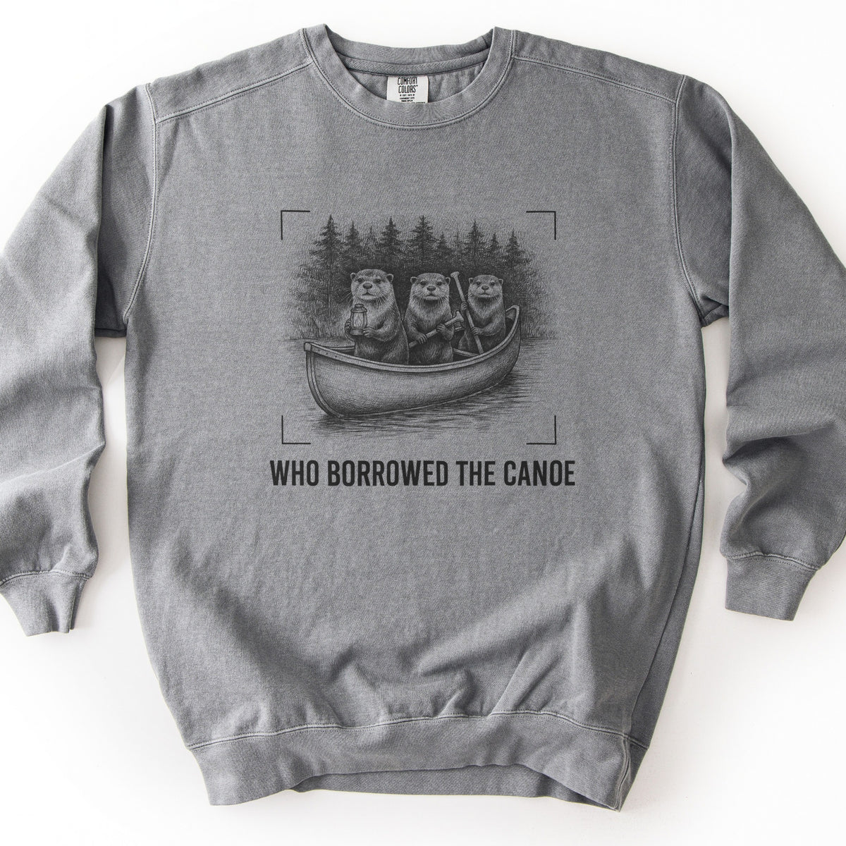 Who Borrowed The Canoe Comfort Colors crewneck sweatshirt in Grey, laid flat. A black sketch of three otters in a canoe, one holding a lantern and two holding paddles, with pine trees behind and corner brackets around it, above the words WHO BORROWED THE CANOE. Laid on white.