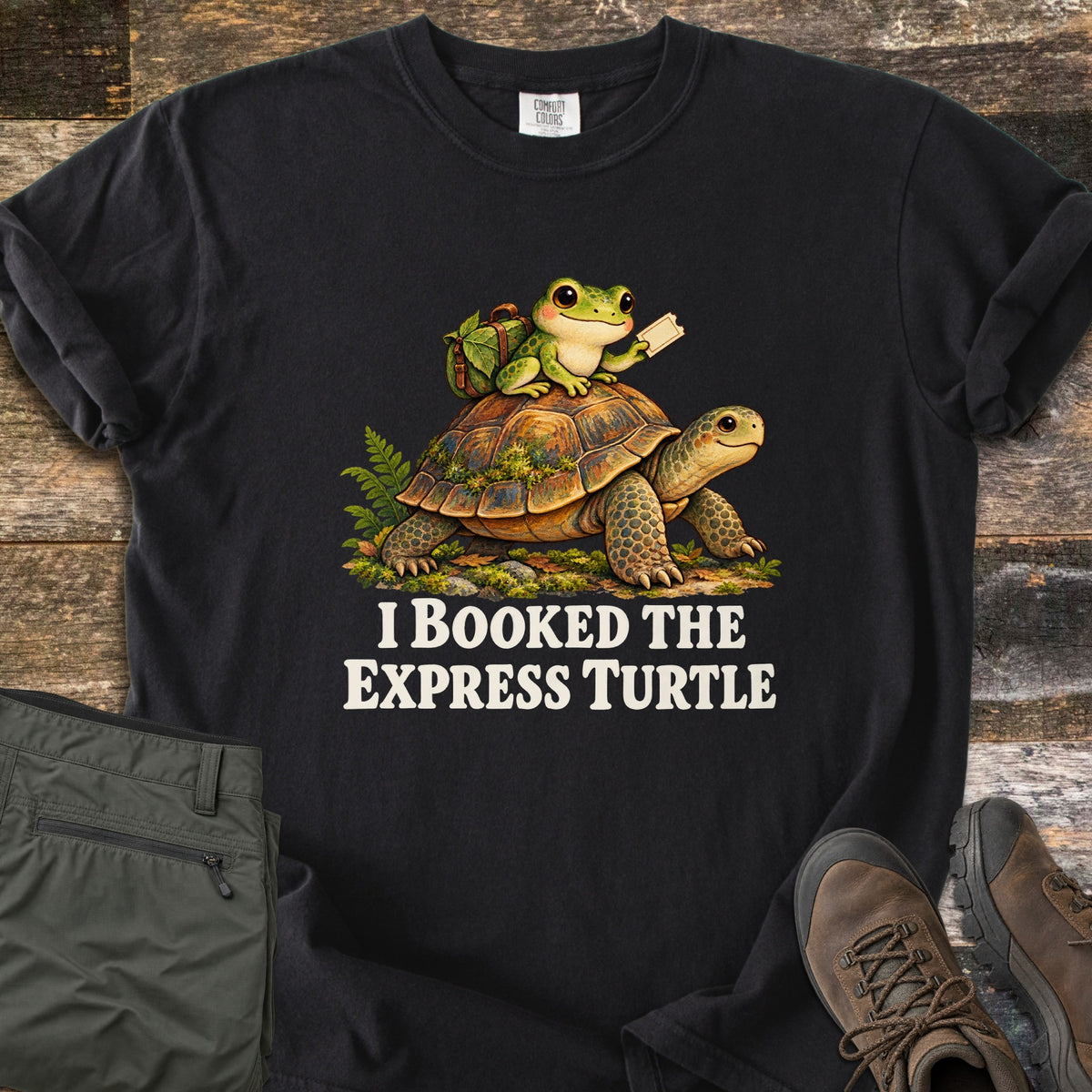 I Booked the Express Turtle Comfort Colors T-shirt in Black, laid flat. A cartoon frog with a leafy backpack holds a ticket while riding a mossy tortoise, above the words I Booked the Express Turtle in cream. Shown on weathered wood with folded olive pants and brown hiking boots.