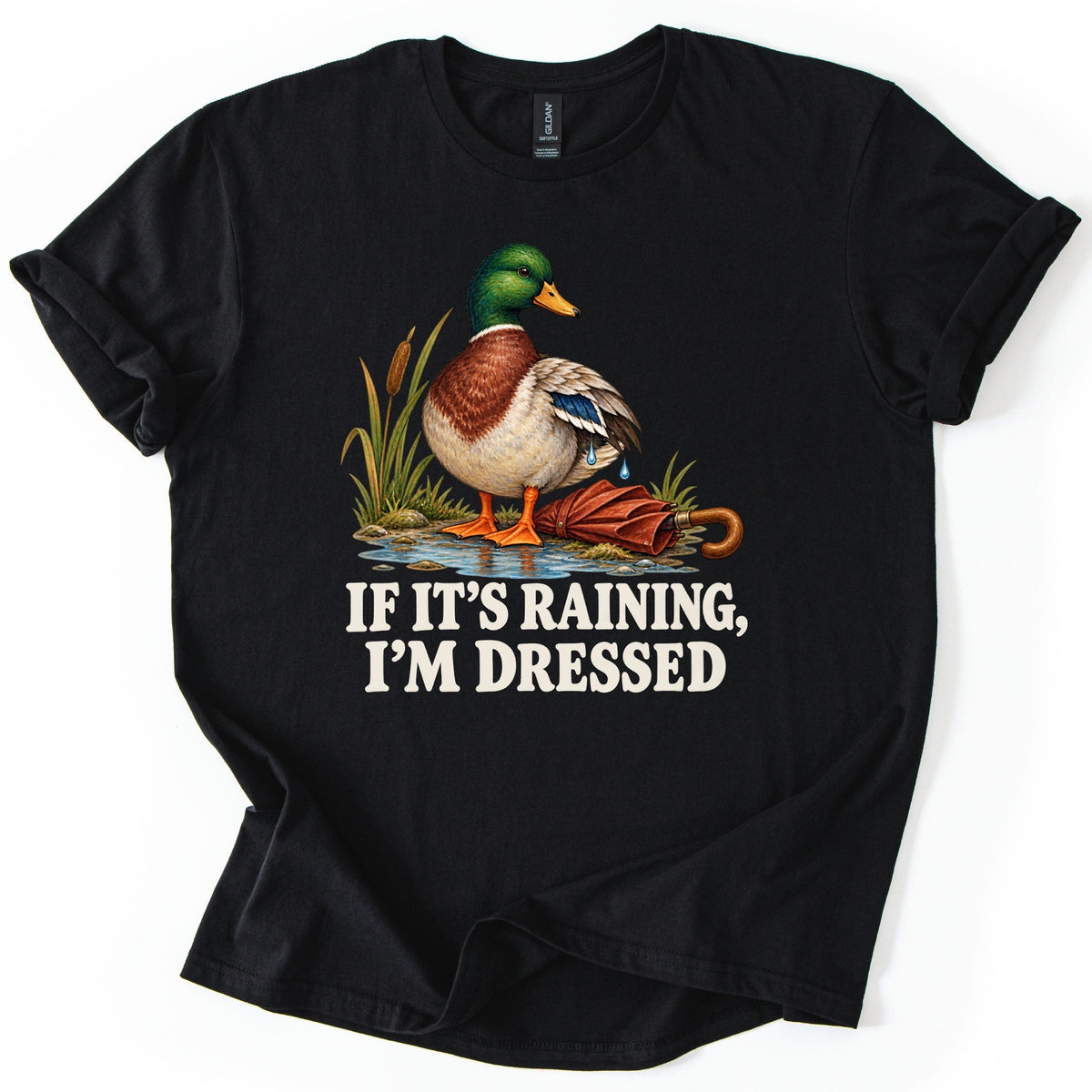 If It's Raining, I'm Dressed Comfort Colors T-shirt in Black, laid flat. A mallard duck with water dripping off its wing stands in a puddle by cattails beside a closed red umbrella, above the words IF IT'S RAINING, I'M DRESSED in cream. Cuffed sleeves, white background.