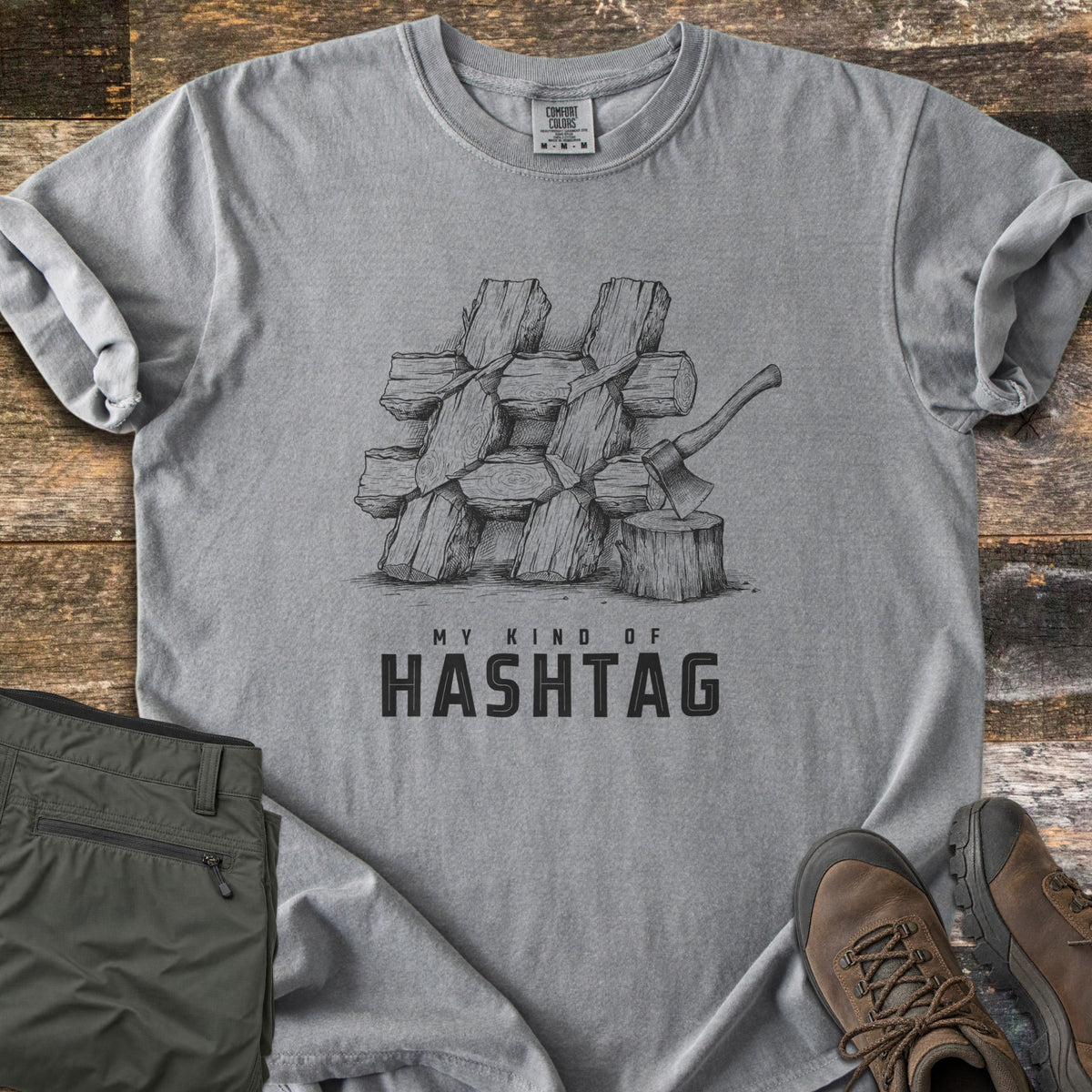 My Kind Of Hashtag Comfort Colors T-shirt in Grey, laid flat. A sketched stack of split firewood logs arranged in a hashtag shape, with an axe in a stump, above the words My Kind Of Hashtag. Olive hiking pants and brown hiking boots lie beside it on weathered wood planks.
