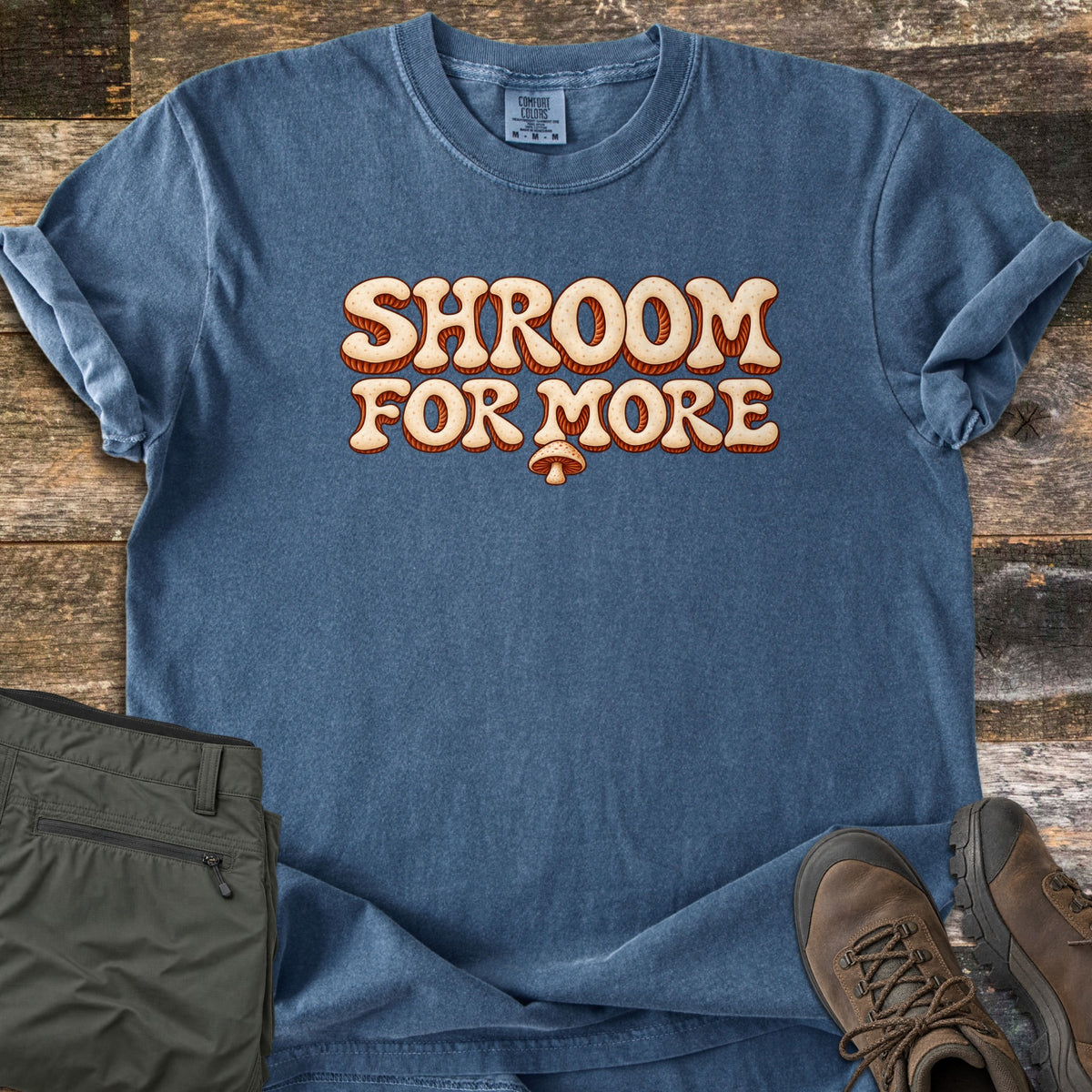 Shroom for More Comfort Colors T-shirt in Denim, laid flat. The words Shroom for More in puffy, retro cream letters with rust-orange shading and tiny speckles, with a small mushroom below. Olive cargo shorts and brown hiking boots sit on weathered wood planks.