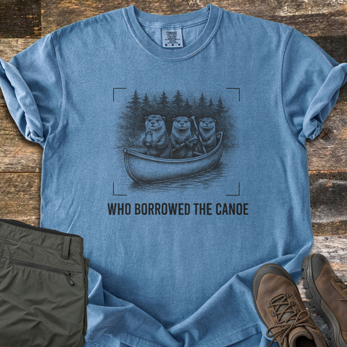 Who Borrowed The Canoe Comfort Colors T-shirt in Blue Jean, laid flat. A black sketch of three otters in a canoe on a lake with pine trees behind, one holding a lantern, framed by camera corner marks above the words Who Borrowed The Canoe. Olive pants and brown boots sit on wood.