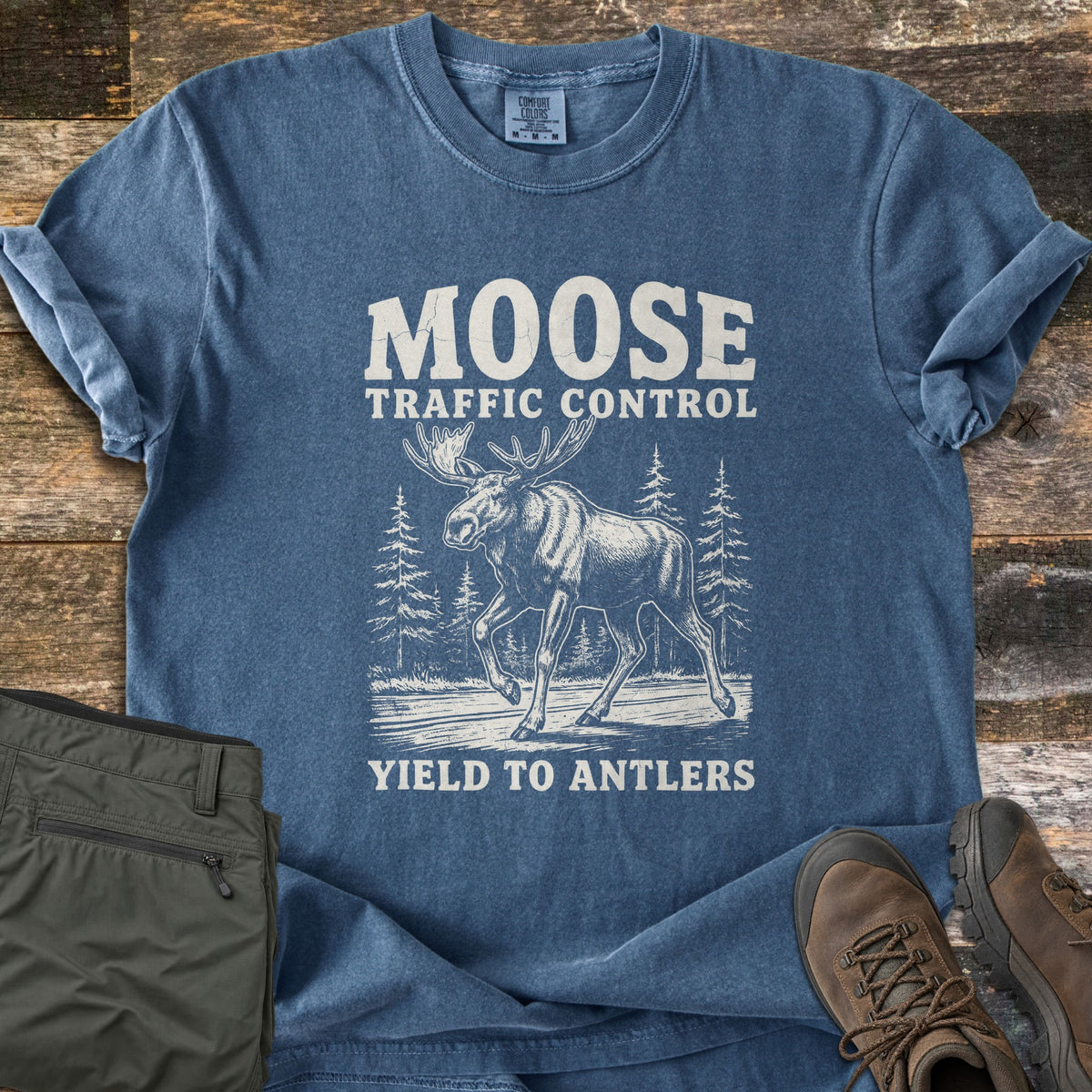 Moose Traffic Control Comfort Colors T-shirt in Denim, laid flat. A vintage-style cream print shows a moose walking across a road among pine trees, with the words Moose Traffic Control above and Yield to Antlers below. Olive pants and brown hiking boots lie beside it on rustic wood.