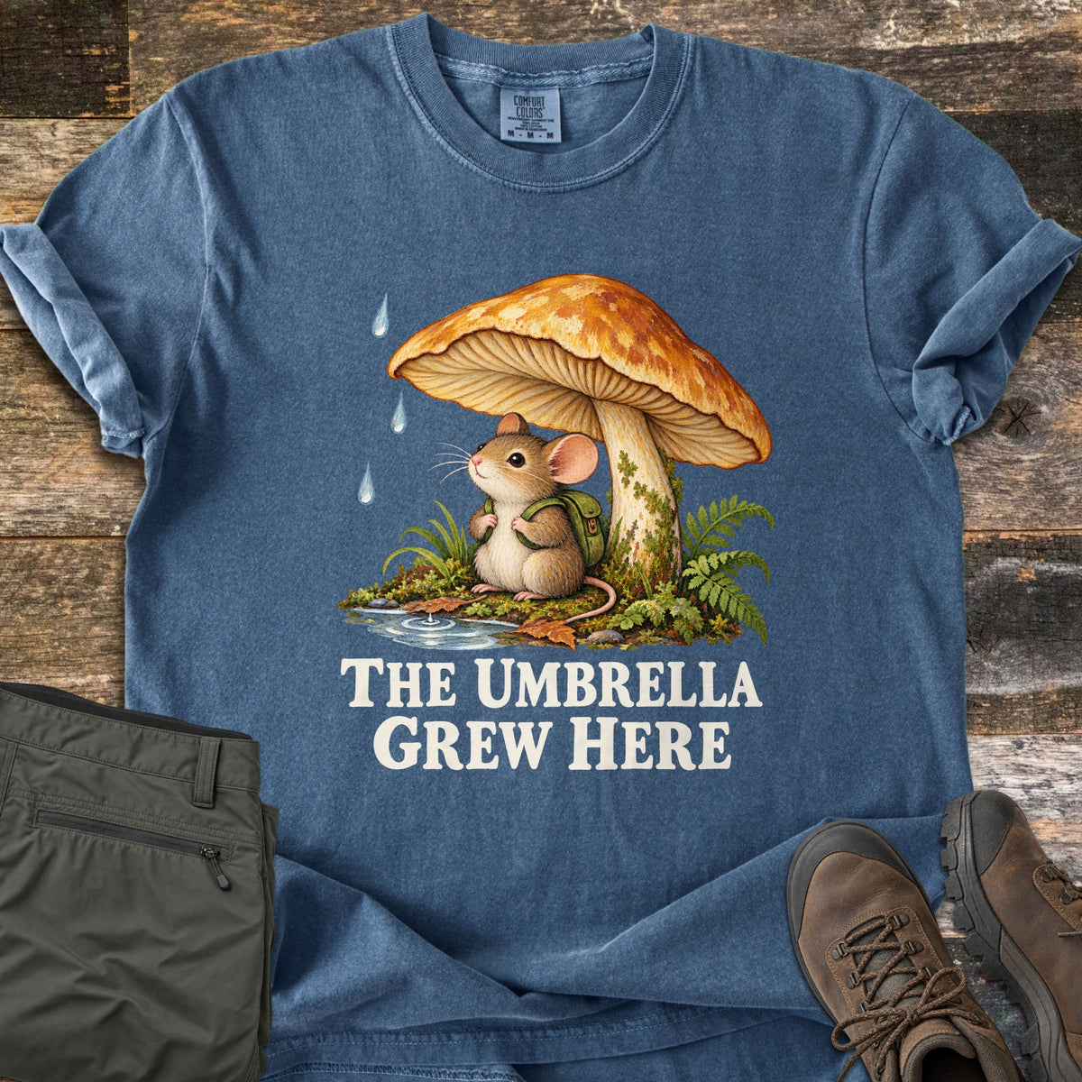 The Umbrella Grew Here Comfort Colors T-shirt in Denim, laid flat. A mouse with a green backpack shelters from raindrops under a large orange mushroom amid ferns and moss, above the words The Umbrella Grew Here. Olive hiking pants and brown boots lie beside it on weathered wood.