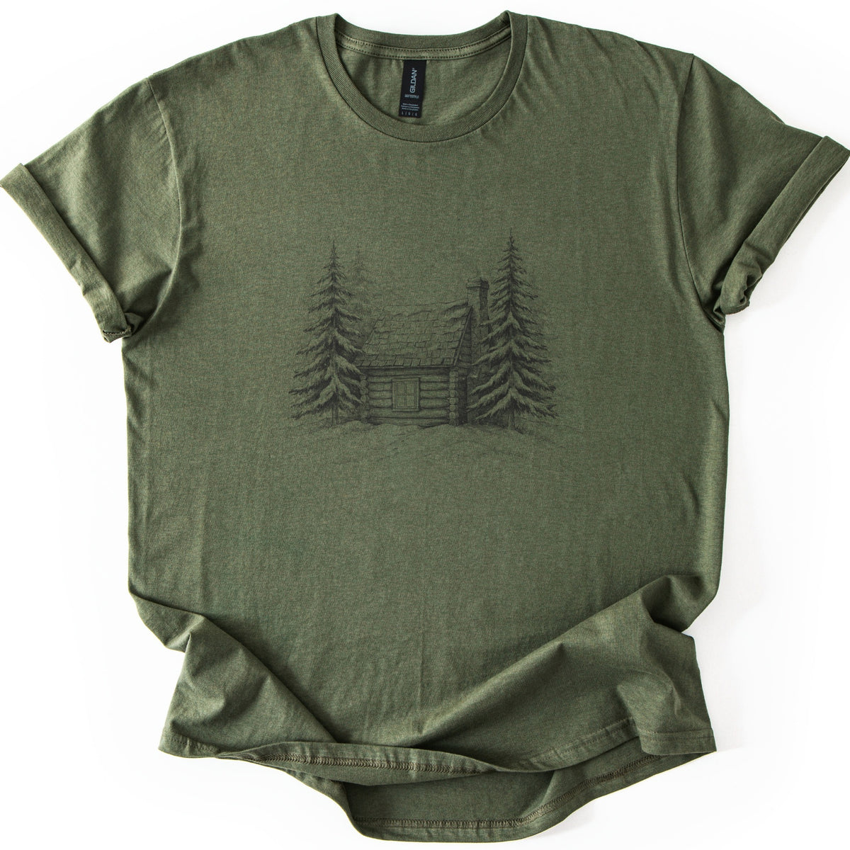 Snowy Log Cabin Comfort Colors T-shirt in Military Green, laid flat. A dark pencil-style sketch of a log cabin with a chimney, flanked by tall pine trees on a snowy drift. No words. Sleeves are rolled on a white background.