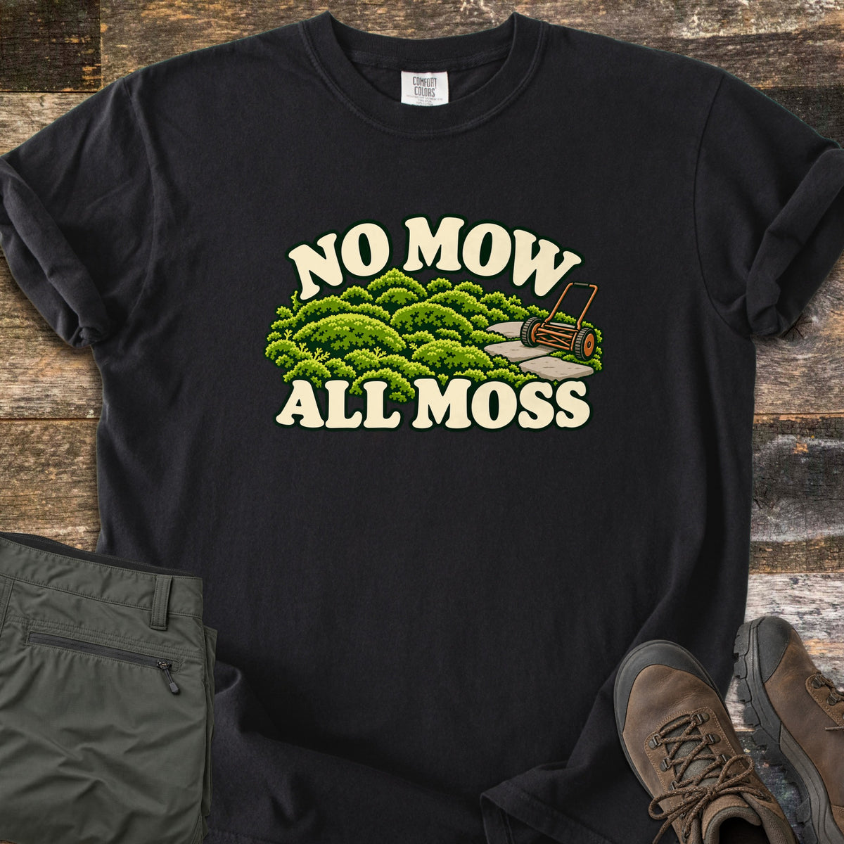 No Mow All Moss Comfort Colors T-shirt in Black, laid flat. Green moss mounds, a stone path and an orange reel mower are printed between the cream words No Mow All Moss. Sleeves are rolled on a weathered wood floor beside folded olive cargo pants and brown hiking boots.