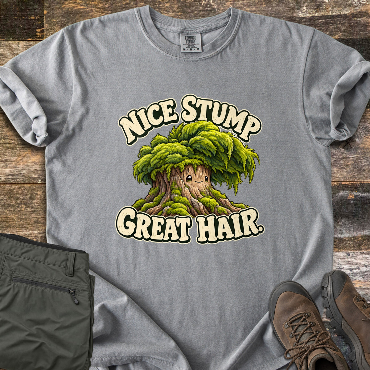 Nice Stump Great Hair Comfort Colors T-shirt in Grey, laid flat. A smiling cartoon tree stump with a shaggy mop of green moss hair sits between the words Nice Stump Great Hair. Sleeves rolled, on weathered wood with folded olive hiking pants and brown hiking boots.