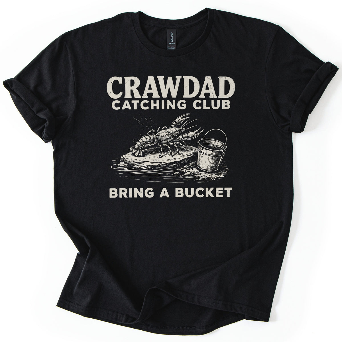 Crawdad Catching Club Comfort Colors T-shirt in Black, laid flat. Cream print reads Crawdad Catching Club above a sketched crawdad on a flat rock at the water's edge beside a metal bucket, with Bring a Bucket below. Cuffed sleeves on a white background.