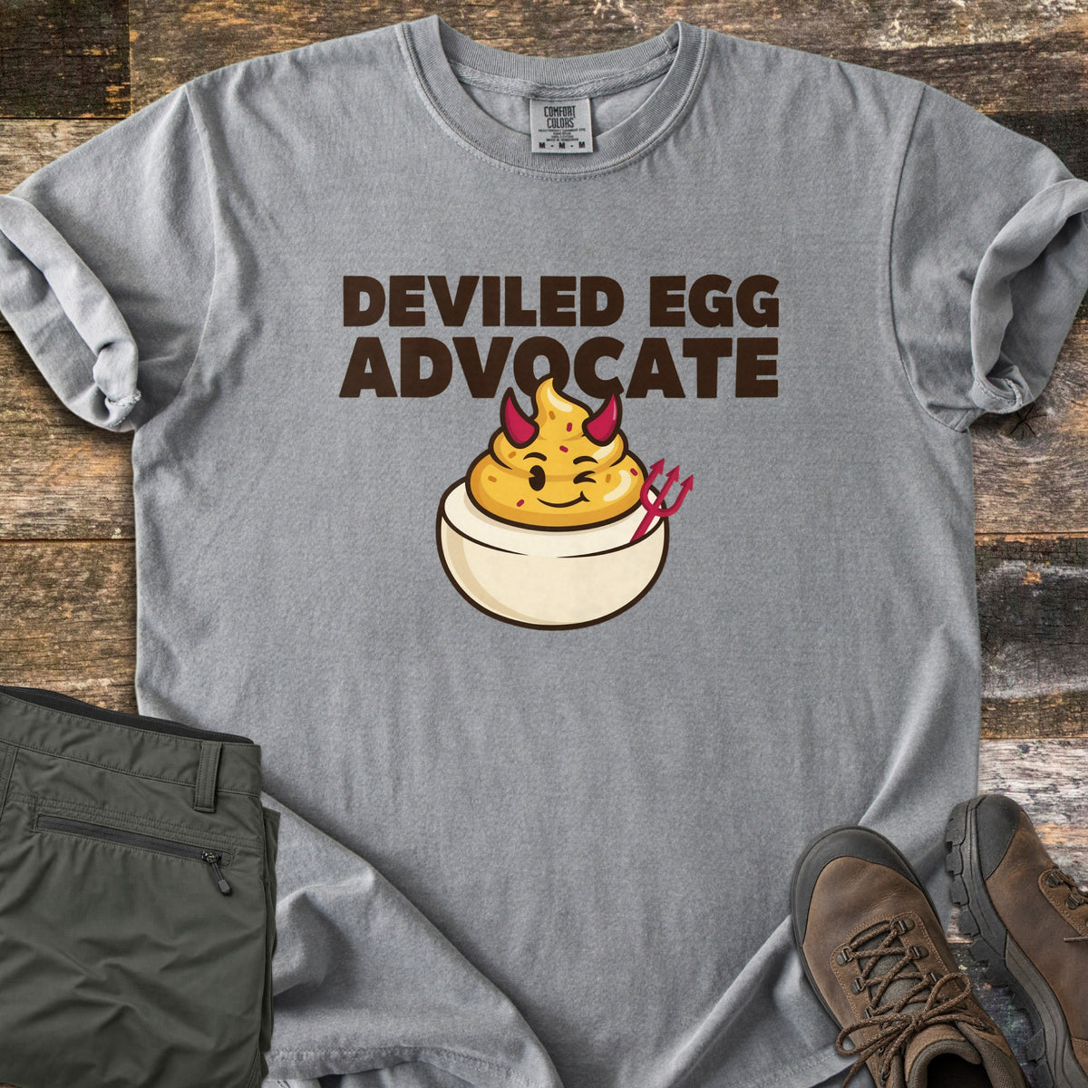 Deviled Egg Advocate Comfort Colors T-shirt in Grey, laid flat. Dark brown letters read Deviled Egg Advocate above a winking cartoon deviled egg with a piped yellow yolk, red devil horns and a red pitchfork. Olive hiking pants and brown boots lie beside it on rustic wood.