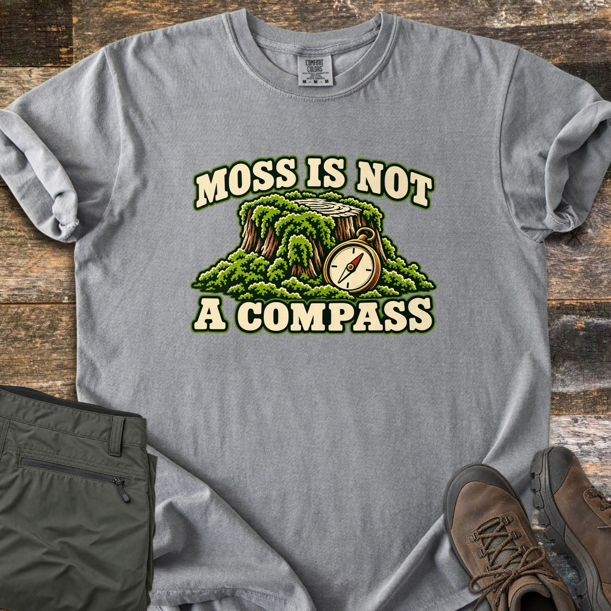 Moss Is Not a Compass Comfort Colors T-shirt in Grey, laid flat. A moss-covered stump with a brass compass at its side appears between the words MOSS IS NOT and A COMPASS. Olive hiking pants and brown leather hiking boots lie on weathered planks.