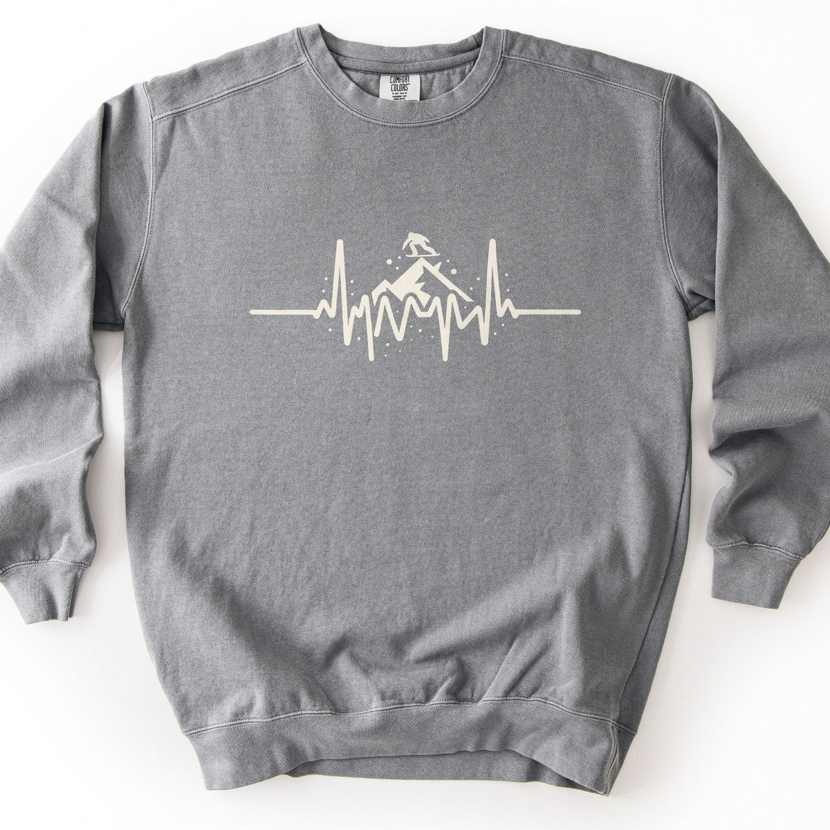 Snow Boarder Heartbeat Comfort Colors crewneck sweatshirt in Grey, laid flat. A cream heartbeat line runs across the chest, rising into a mountain peak with a snowboarder riding on top and small dots of snow around it. No words. The sweatshirt lies on a white surface.