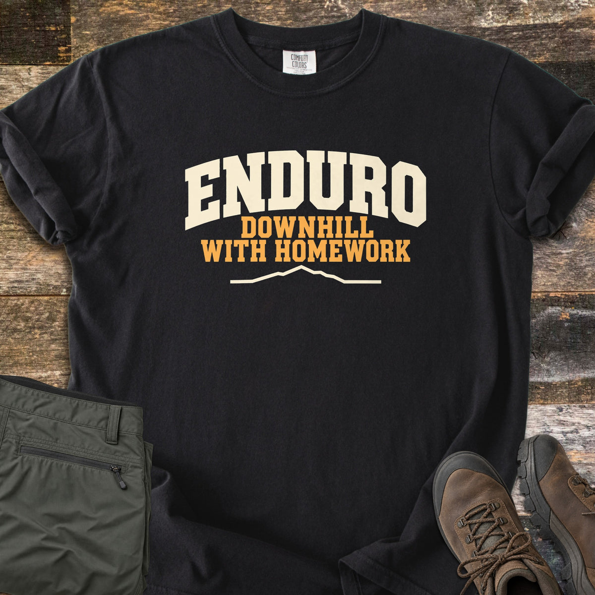 Enduro Downhill With Homework Comfort Colors T-shirt in Black, laid flat. The word Enduro in large cream arched letters, with Downhill With Homework in orange below and a thin cream mountain ridge line. Sleeves are rolled on a rustic wood surface beside green pants and brown hiking boots.