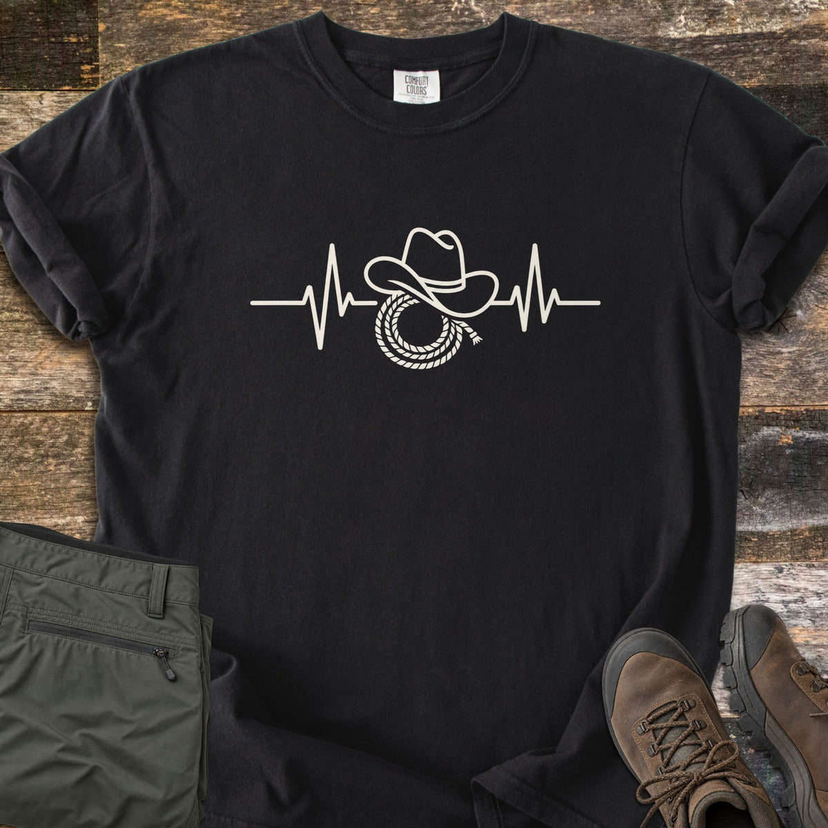 Cowboy Heartbeat Comfort Colors T-shirt in Black, laid flat. A cream line art heartbeat line rises into peaks around a cowboy hat resting on a coiled lasso; there are no words. Sleeves are cuffed, on rustic wood boards beside folded olive cargo pants and brown hiking boots.