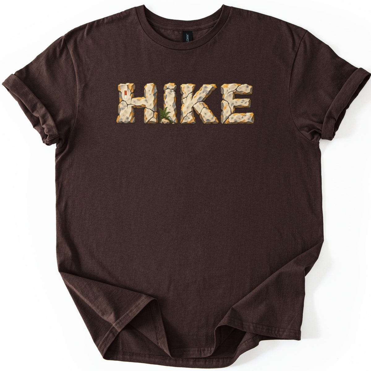 Hike Comfort Colors T-shirt in Dark Chocolate, laid flat. The word HIKE is printed in letters made of cracked tan stone with orange edges, a small green fern at the base and a red and white trail marker on the H. Sleeves rolled, on a white background.