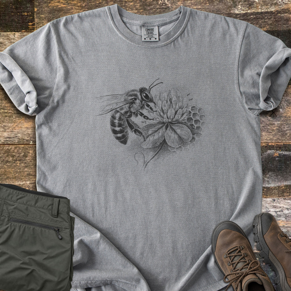 Honeybee On Clover Comfort Colors T-shirt in Grey, laid flat. A detailed black engraving-style illustration of a honeybee on a clover blossom with honeycomb cells behind it, no words. Sleeves rolled, on weathered wood planks beside olive hiking pants and brown hiking boots.
