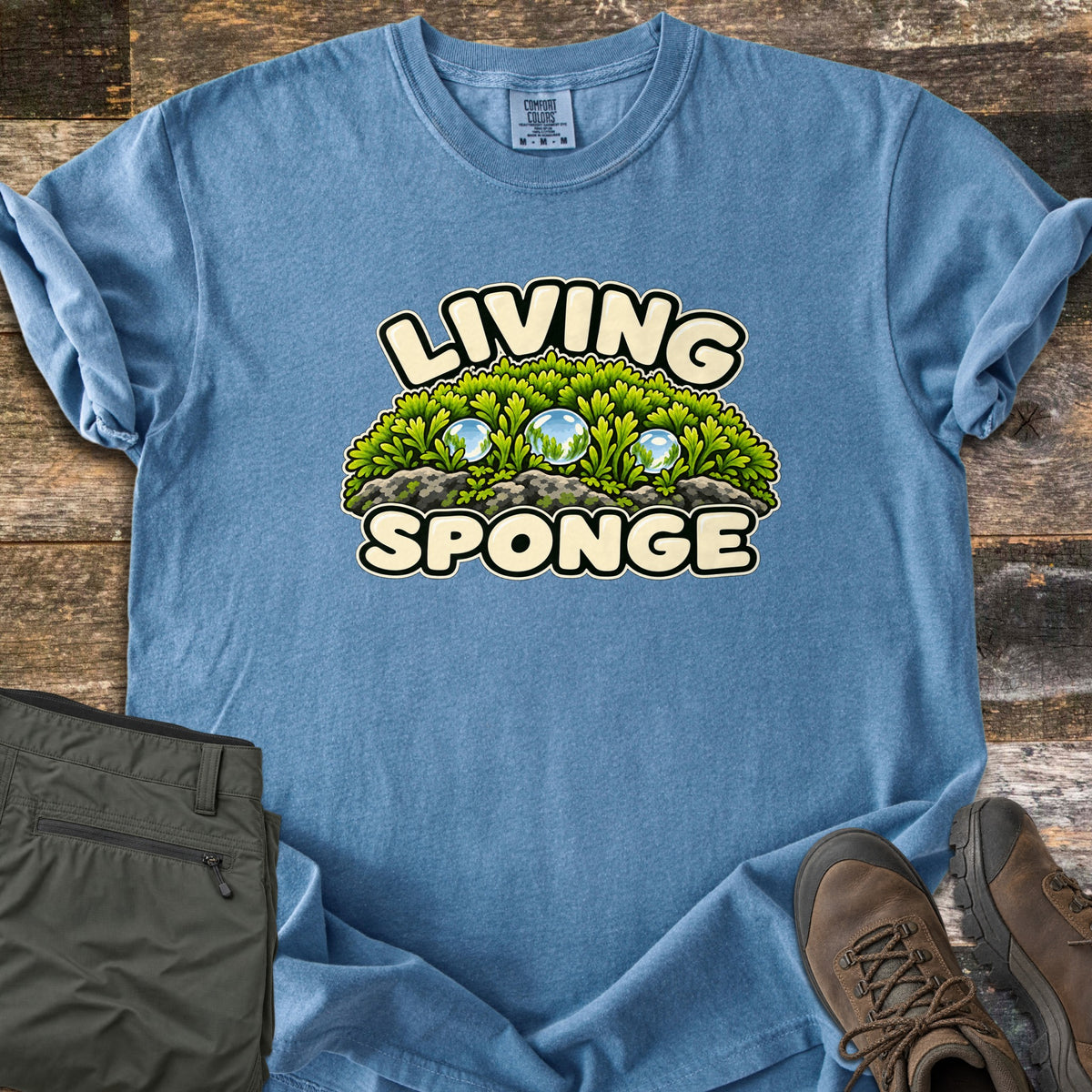 Living Sponge Comfort Colors T-shirt in Blue Jean, laid flat. A cartoon mound of bright green moss on gray rock, holding three clear water droplets, between the words Living Sponge. Olive hiking pants and brown leather hiking boots lie beside it on weathered wood.