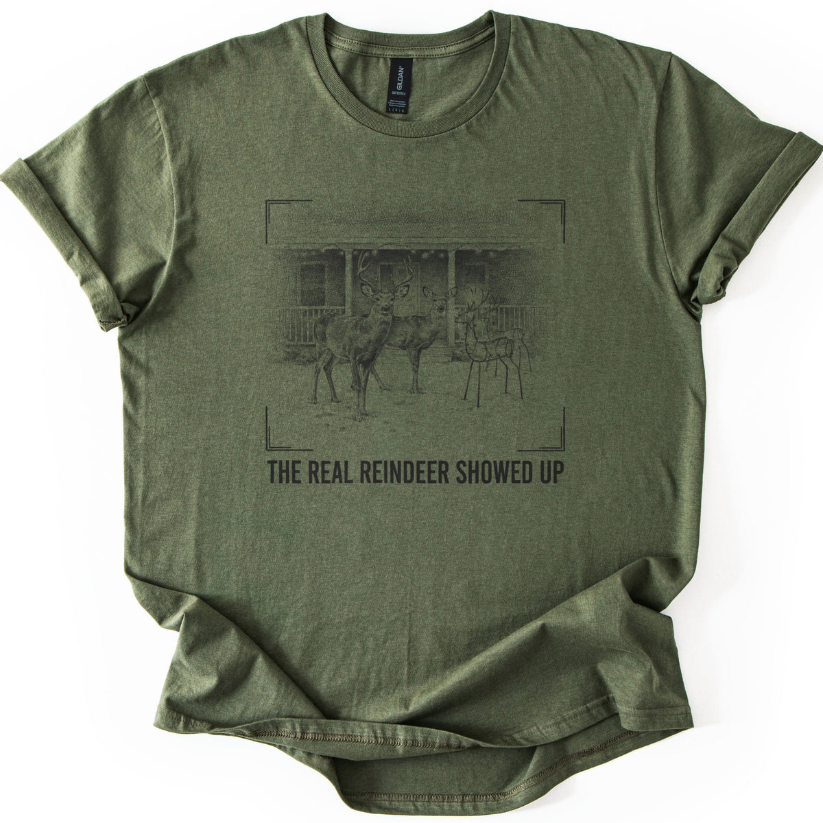 The Real Reindeer Showed Up Comfort Colors T-shirt in Military Green, laid flat. A dark sketch of a buck and doe standing near wire reindeer lawn decorations in front of a porch, framed by viewfinder corners, above the words The Real Reindeer Showed Up. Sleeves cuffed, on a white background.