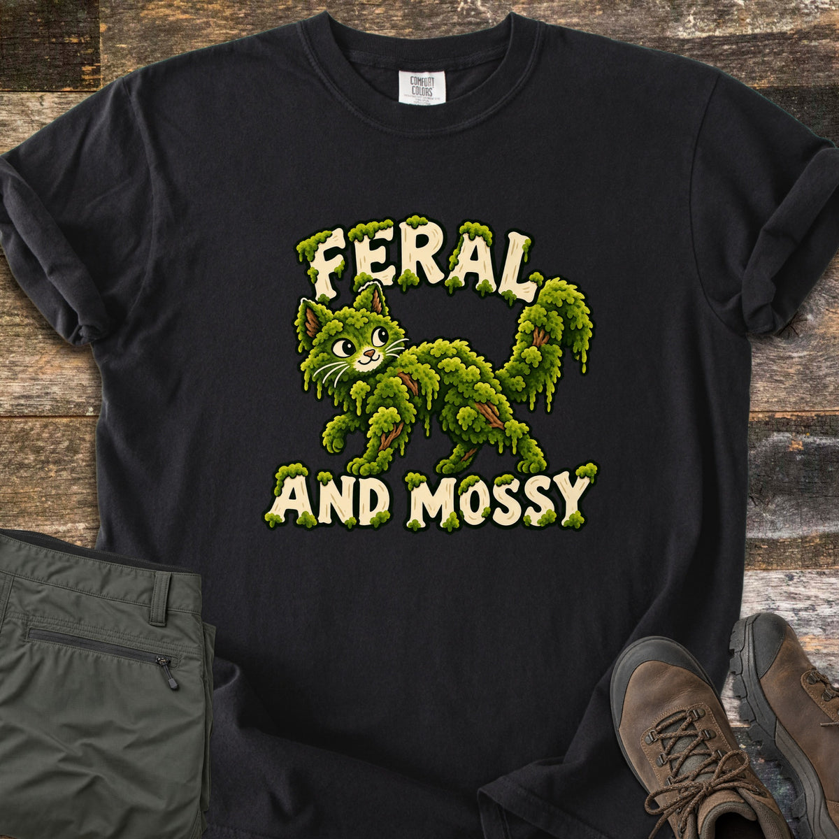 Feral and Mossy Comfort Colors T-shirt in Black, laid flat. A cartoon cat made of green moss with bits of bark walks between the words Feral and Mossy in cream letters dripping with moss. Folded olive pants and brown hiking boots sit on a weathered wood floor.