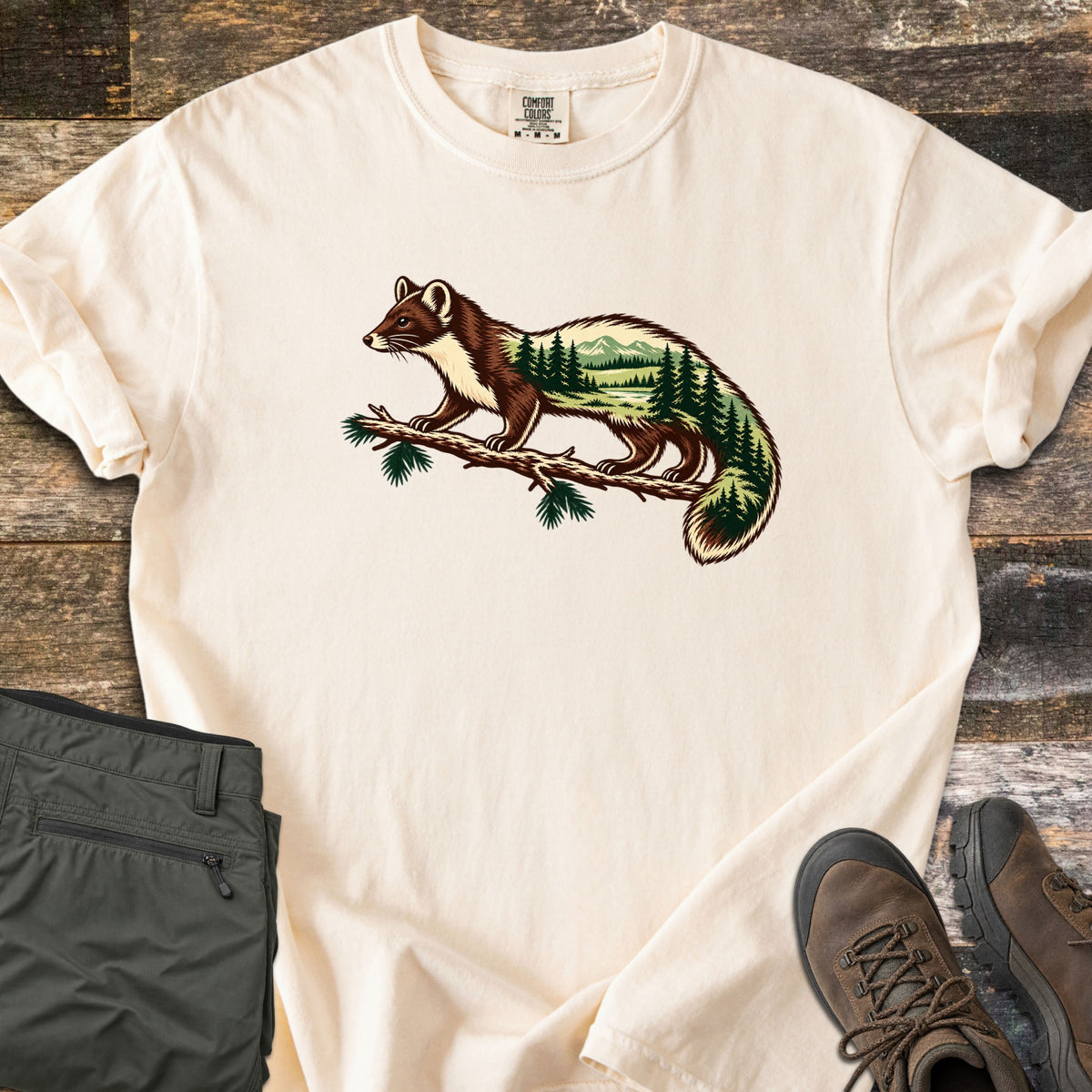 Pine Marten Forest Comfort Colors T-shirt in Ivory, laid flat. A pine marten stands on a pine branch, its body filled with a scene of evergreen trees, a lake and mountains. No words are printed. Shown on weathered wood with folded olive pants and brown hiking boots.