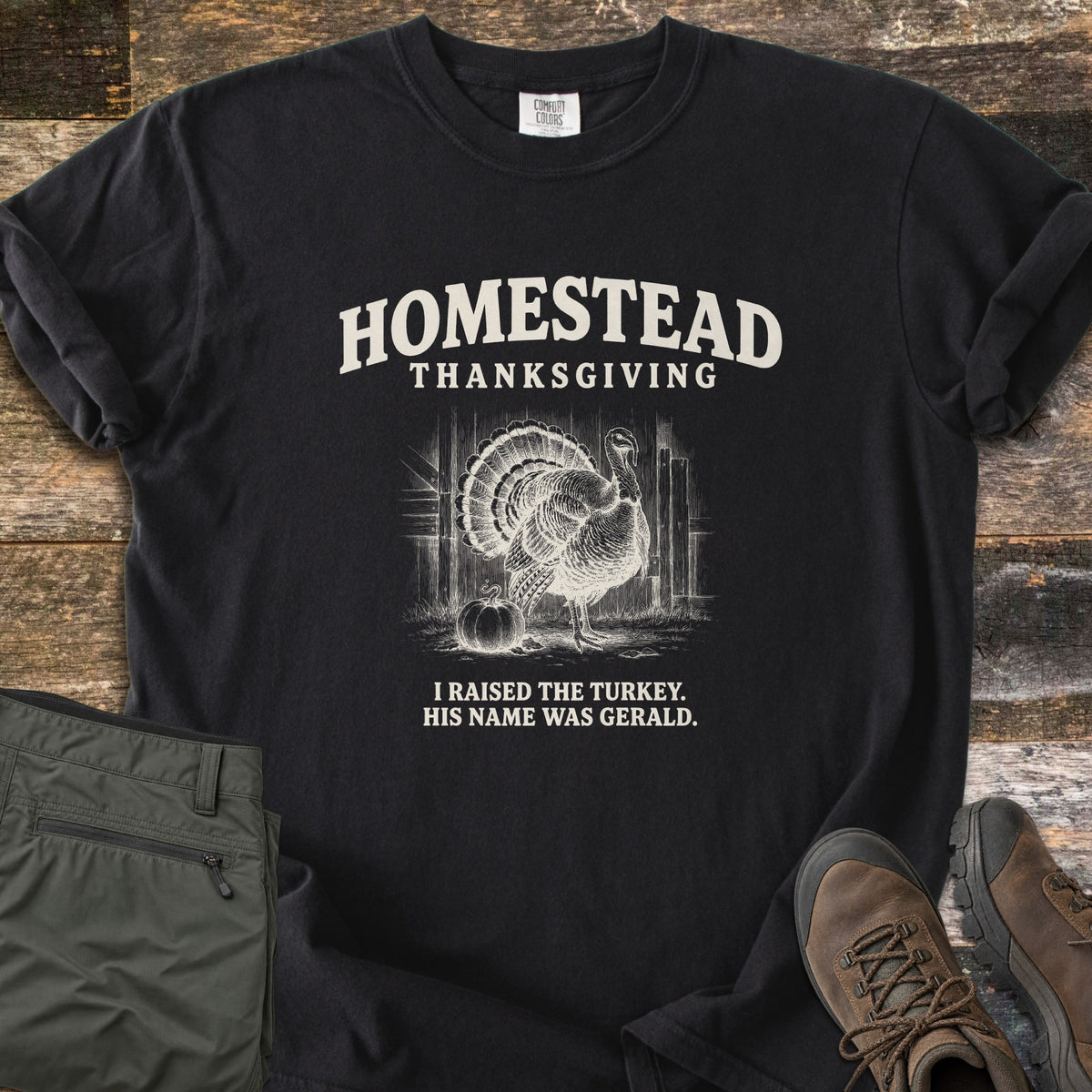 Homestead Thanksgiving Comfort Colors T-shirt in Black, laid flat. Cream print of a fanned-tail turkey by a barn door and pumpkin, under the words Homestead Thanksgiving, above I Raised the Turkey. His Name Was Gerald. On weathered wood with olive pants and brown hiking boots.