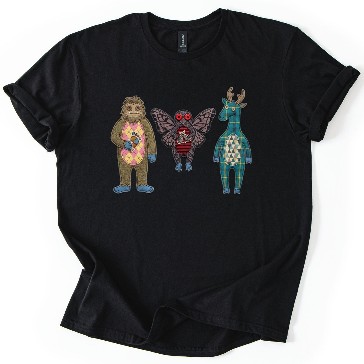 Patchwork Cryptid Trio Comfort Colors T-shirt in Black, laid flat. Three fabric patchwork cryptids with no text: Bigfoot in brown felt with a pink argyle belly, Mothman with red button eyes, and a teal plaid antlered creature with a triangle belly. On white.