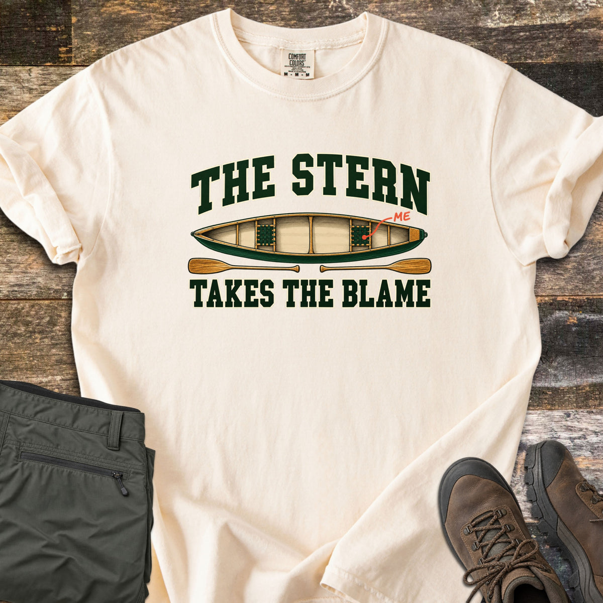 The Stern Takes the Blame Comfort Colors T-shirt in Ivory, laid flat. Dark green varsity letters read The Stern above a top-down canoe whose back seat is marked ME by a red arrow, then two paddles and Takes the Blame. Olive pants and brown hiking boots lie on weathered boards.