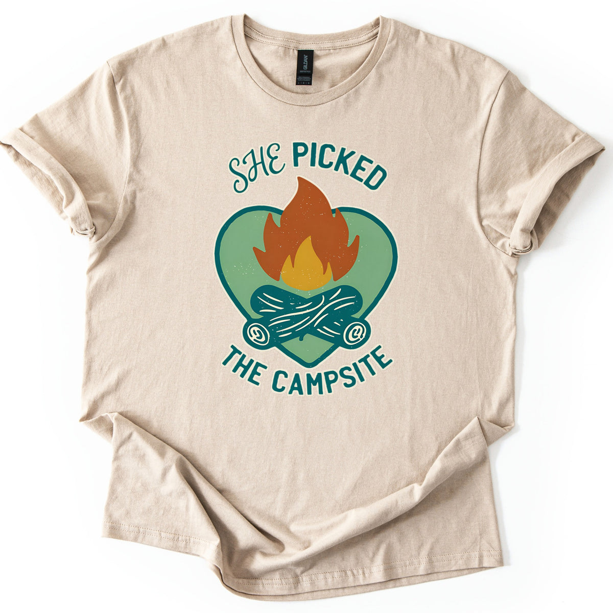 She Picked The Campsite Comfort Colors T-shirt in Sand, laid flat. An orange and yellow campfire on crossed teal logs inside a sage green heart, with the words SHE PICKED above and THE CAMPSITE below. The shirt lies on a white surface with cuffed sleeves and a folded hem.