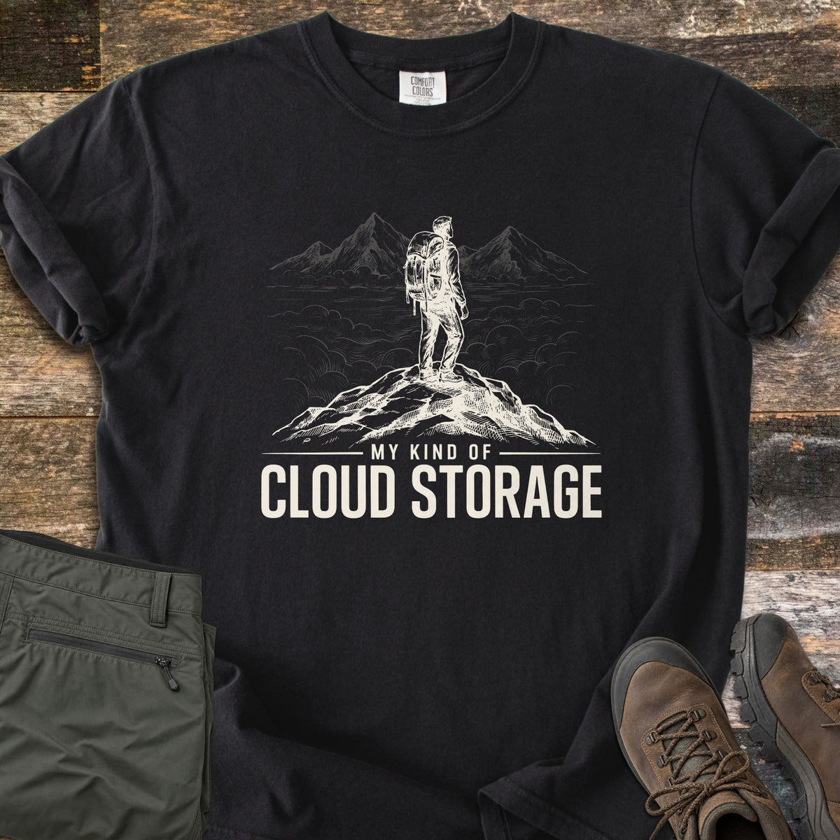 My Kind Of Cloud Storage Comfort Colors T-shirt in Black, laid flat. A cream sketch of a backpacker on a rocky summit facing peaks above the clouds, over the words My Kind Of Cloud Storage. Sleeves are rolled, with olive hiking pants and brown hiking boots beside it on weathered wood.