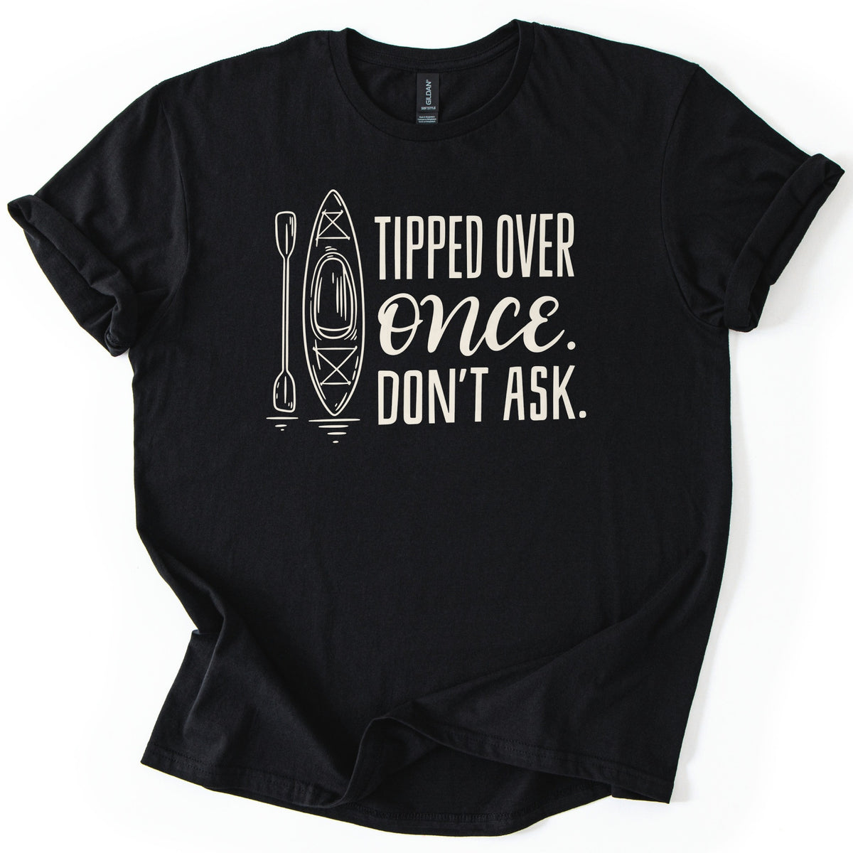 Tipped Over Once Kayak Comfort Colors T-shirt in Black, laid flat. A cream line drawing of an upright kayak and paddle with water ripples sits beside the words Tipped Over Once. Don't Ask., with Once in script. The shirt has cuffed sleeves on a white background.