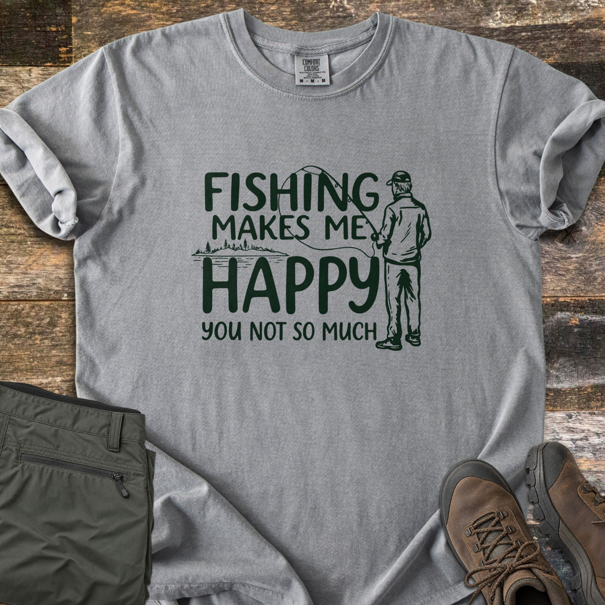 Fishing Makes Me Happy Comfort Colors T-shirt in Grey, laid flat. Dark green print of a man in a cap casting a fishing rod by a tree-lined lake, with the words Fishing Makes Me Happy You Not So Much. Laid on weathered wood with olive pants and brown hiking boots.