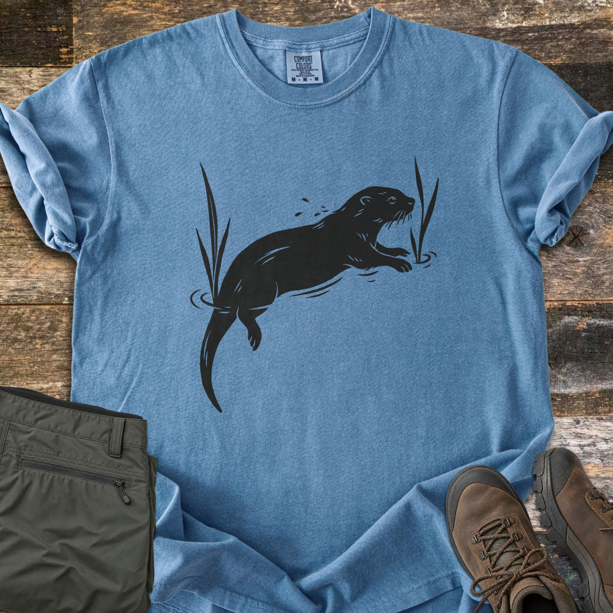 Swimming Otter Comfort Colors T-shirt in Blue Jean, laid flat. A black print of an otter swimming between tall reeds, with ripples and splashing droplets. Sleeves are rolled; olive hiking pants and brown hiking boots sit on weathered wood planks.