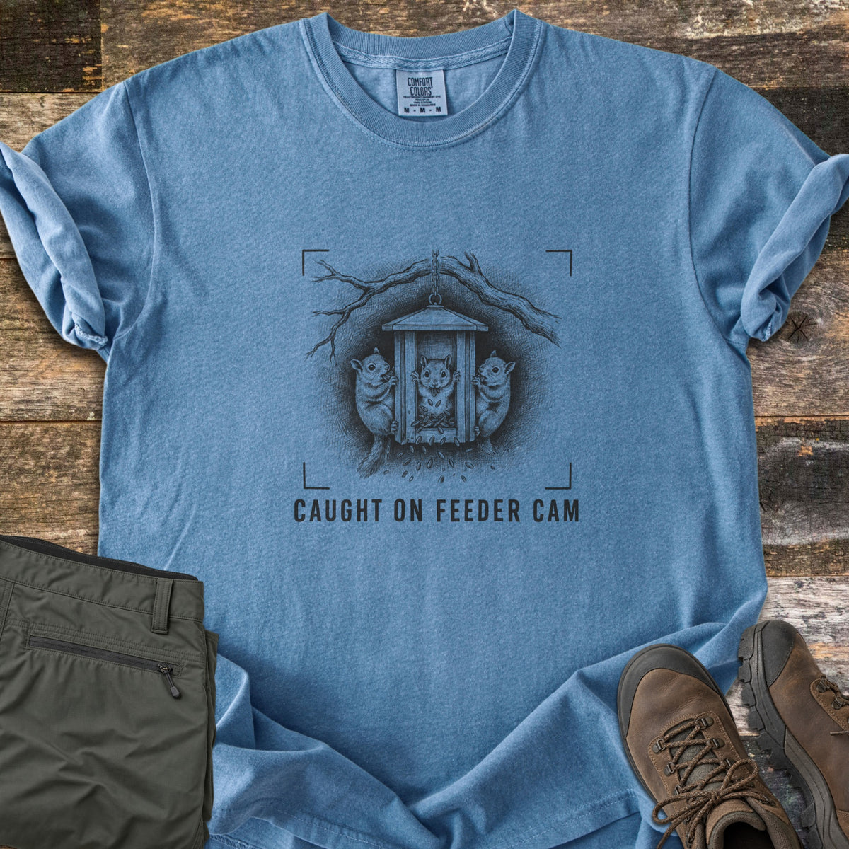 Caught On Feeder Cam Comfort Colors T-shirt in Blue Jean, laid flat. A black ink sketch of three squirrels raiding a hanging wooden feeder inside camera viewfinder corners, above the words CAUGHT ON FEEDER CAM. Folded olive pants and brown hiking boots sit on weathered wood planks.