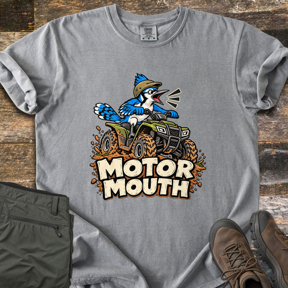 Motor Mouth Comfort Colors T-shirt in Grey, laid flat. A cartoon blue jay wearing a tan helmet squawks from a green ATV spraying mud, above the words Motor Mouth. Folded olive pants and brown leather hiking boots lie on dark wood planks.