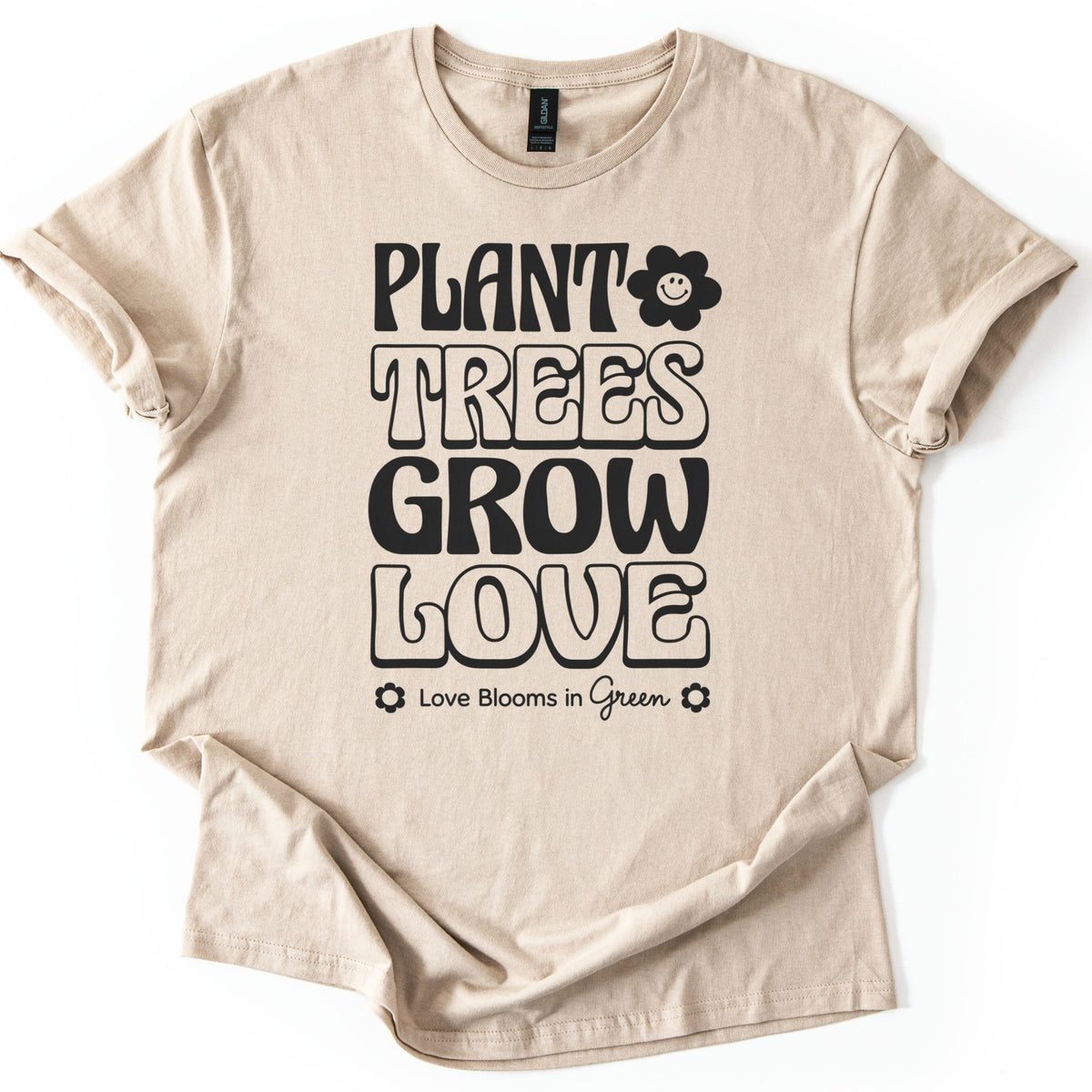 Plant Trees Grow Love Comfort Colors T-shirt in Sand, laid flat. Black retro bubble letters read Plant Trees Grow Love, with a smiling daisy beside Plant and Love Blooms in Green between two small flowers below. Sleeves are rolled on a white background.