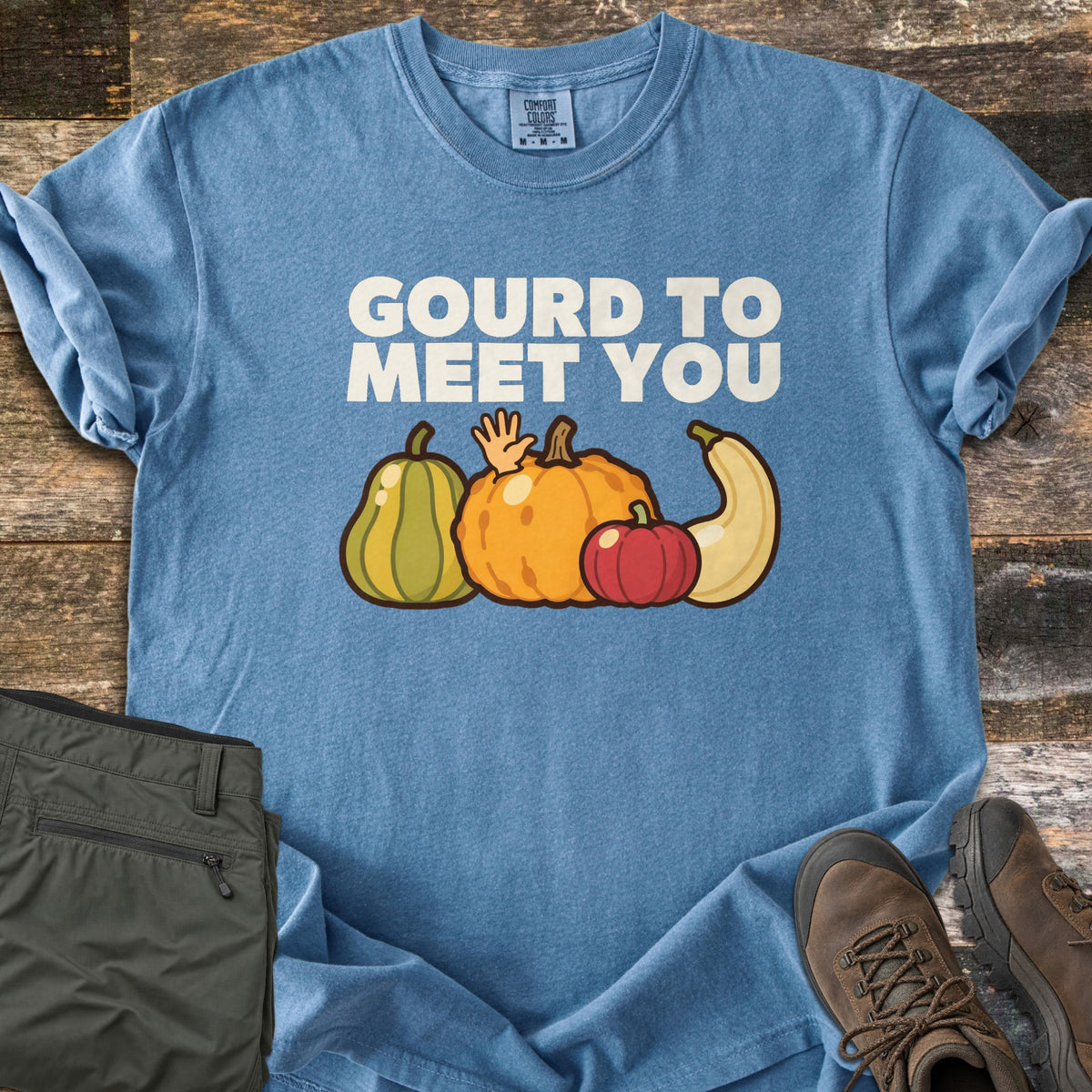 Gourd To Meet You Comfort Colors T-shirt in Blue Jean, laid flat. Off-white words Gourd To Meet You above cartoon gourds: a green striped squash, an orange pumpkin with a waving hand, a red pumpkin and a cream squash. Olive pants and brown hiking boots sit on wood planks.
