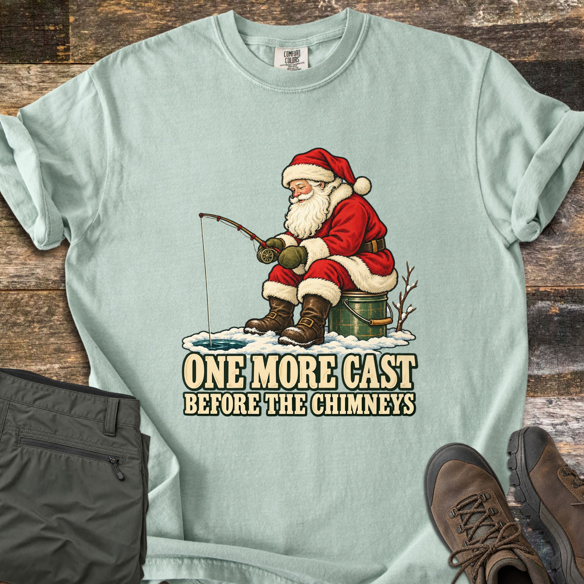 One More Cast Before the Chimneys Comfort Colors T-shirt in Bay, laid flat. Santa in his red suit sits on a green bucket, ice fishing beside a bare branch, above the words One More Cast Before the Chimneys. Olive pants and brown hiking boots lie beside it on weathered wood.