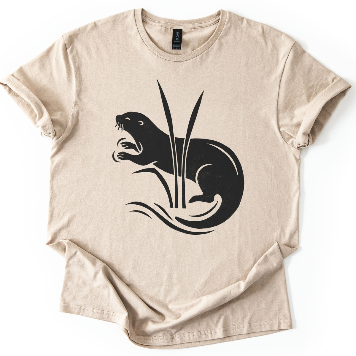 Otter In The Reeds Comfort Colors T-shirt in Sand, laid flat. A black silhouette of an otter with open mouth, whiskers and raised paws curls behind two tall reeds above swirling water lines. No words. The shirt lies on a white surface with sleeves rolled.