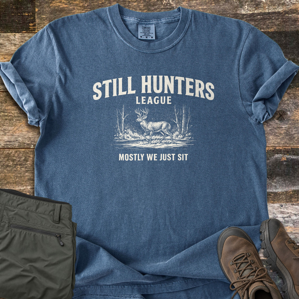 Still Hunters League Comfort Colors T-shirt in Denim, laid flat. Printed in cream: Still Hunters League above a sketched buck standing among bare trees, with Mostly We Just Sit below. Olive cargo pants and brown hiking boots sit beside it on weathered wood planks.