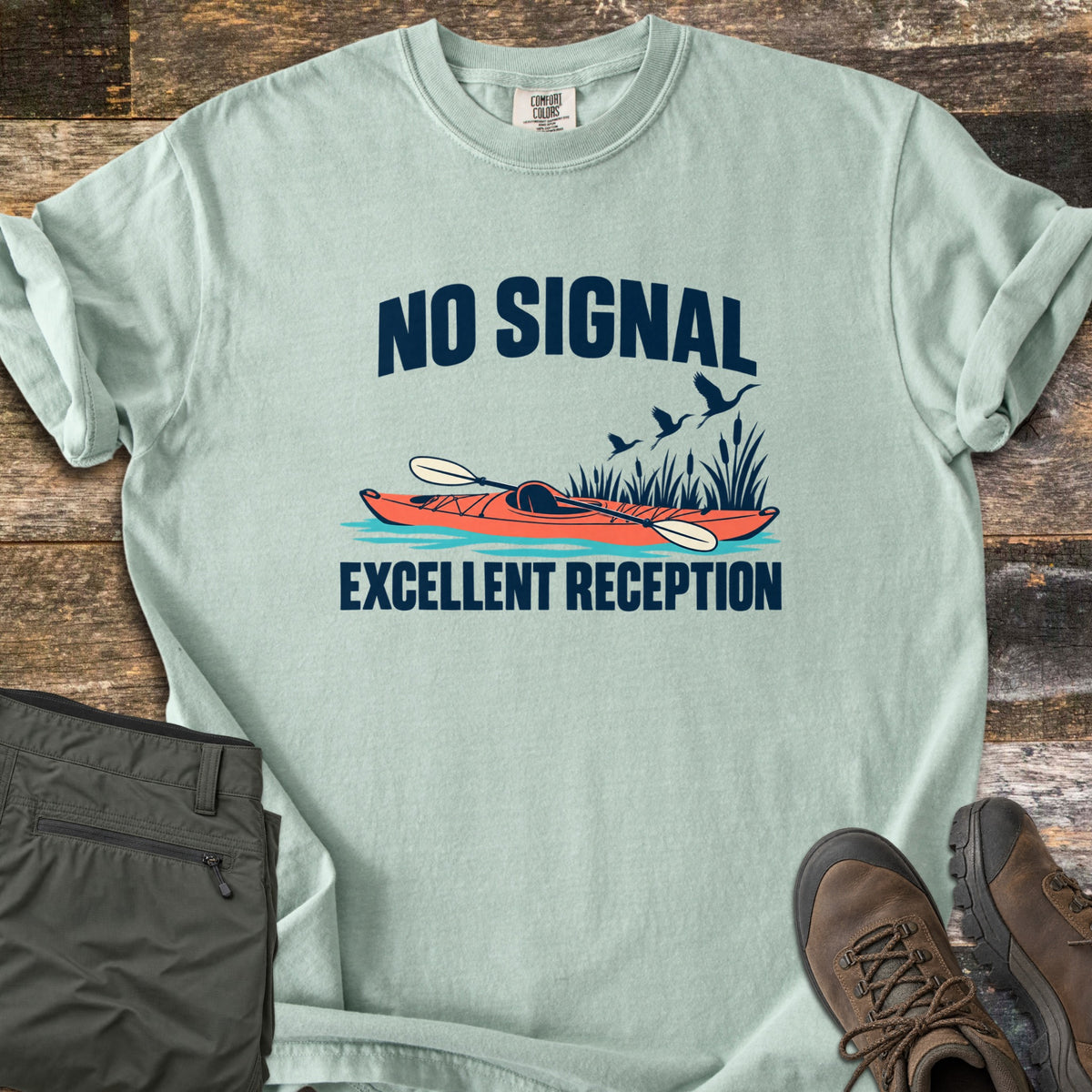 Excellent Reception Comfort Colors T-shirt in Bay, laid flat. The words No Signal arch over an orange kayak with a paddle among reeds and flying birds, above the words Excellent Reception. Olive pants and brown hiking boots sit on a weathered wood surface.