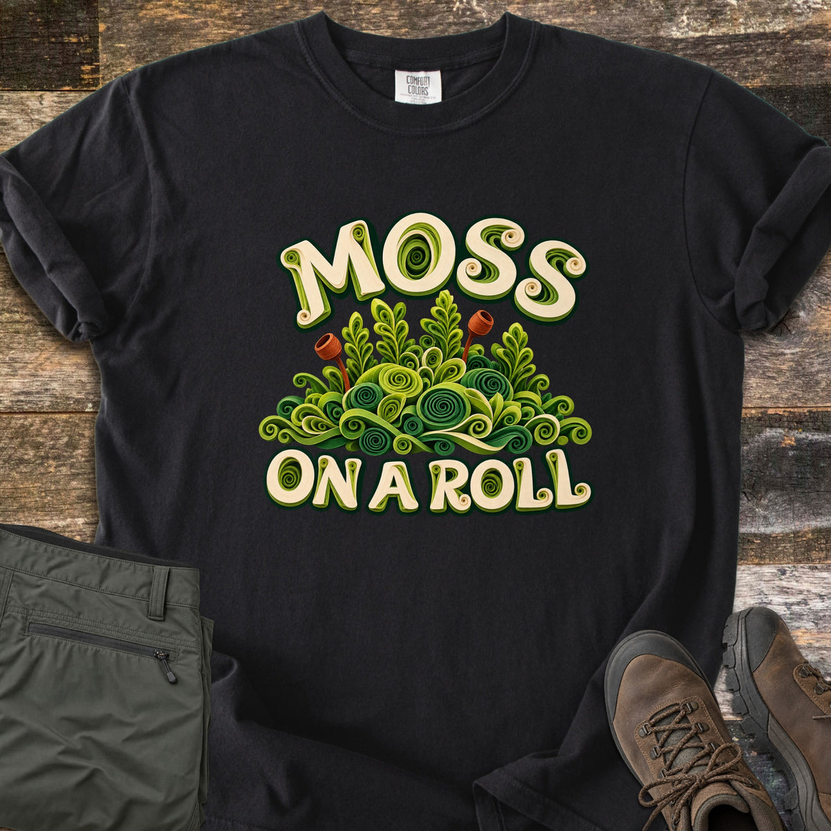 Moss on a Roll Comfort Colors T-shirt in Black, laid flat. Swirled paper-quill style green moss with two rust-colored sprouts sits between the words Moss On A Roll in cream lettering. Sleeves are cuffed on a wood plank floor beside olive hiking pants and brown hiking boots.