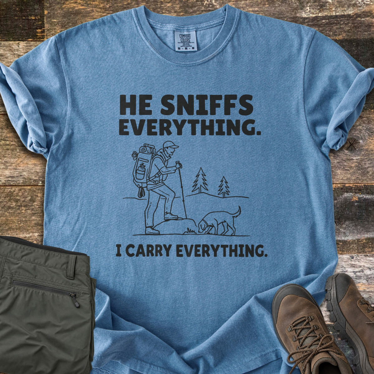 He Sniffs Everything. I Carry Everything Comfort Colors T-shirt in Blue Jean, laid flat. Black line art of a hiker with a large backpack and trekking pole beside a dog sniffing a rock, between the words He Sniffs Everything. and I Carry Everything. Olive pants and brown hiking boots sit on weathered wood.