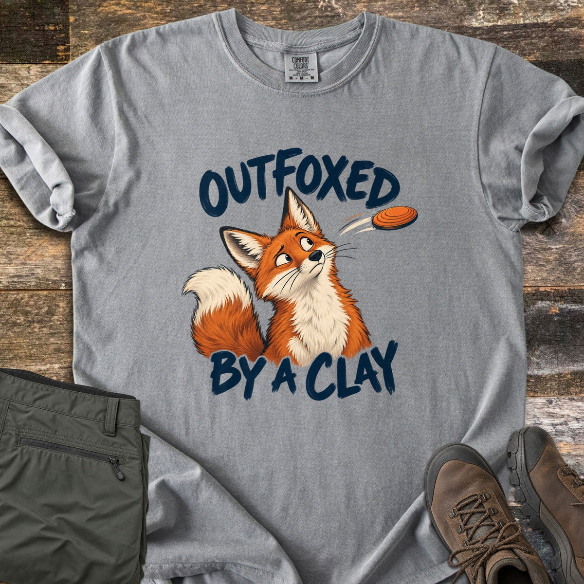 Outfoxed by a Clay Comfort Colors T-shirt in Grey, laid flat. A cartoon red fox glances sideways at an orange clay target flying past, between the words Outfoxed By A Clay in navy brush letters. Sleeves rolled on rustic wood with olive pants and brown hiking boots.
