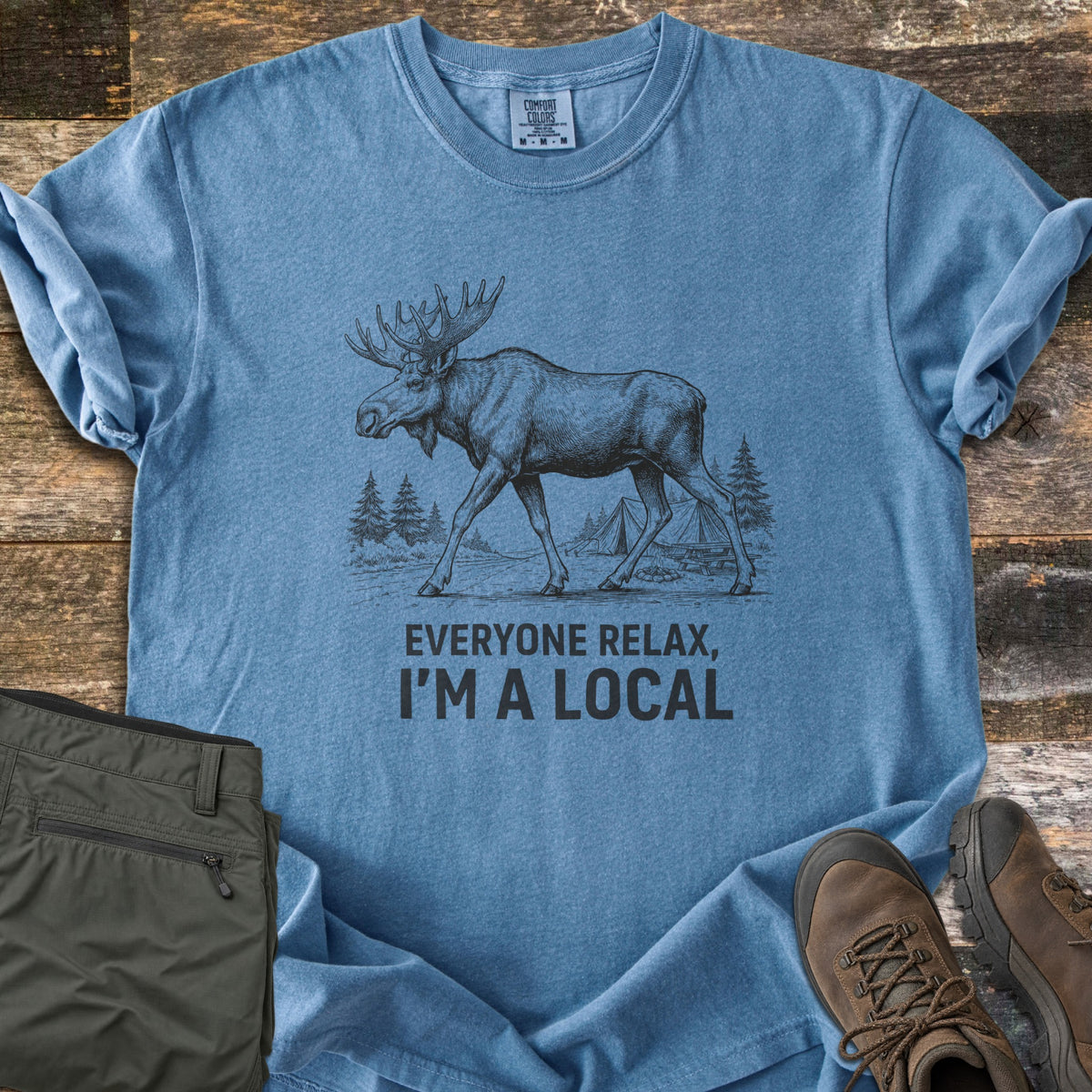 Everyone Relax I'm A Local Comfort Colors T-shirt in Blue Jean, laid flat. A sketched moose walks past pine trees and tents at a campsite above the words Everyone Relax, I'm A Local. Sleeves are rolled on a wood floor beside folded green pants and brown hiking boots.