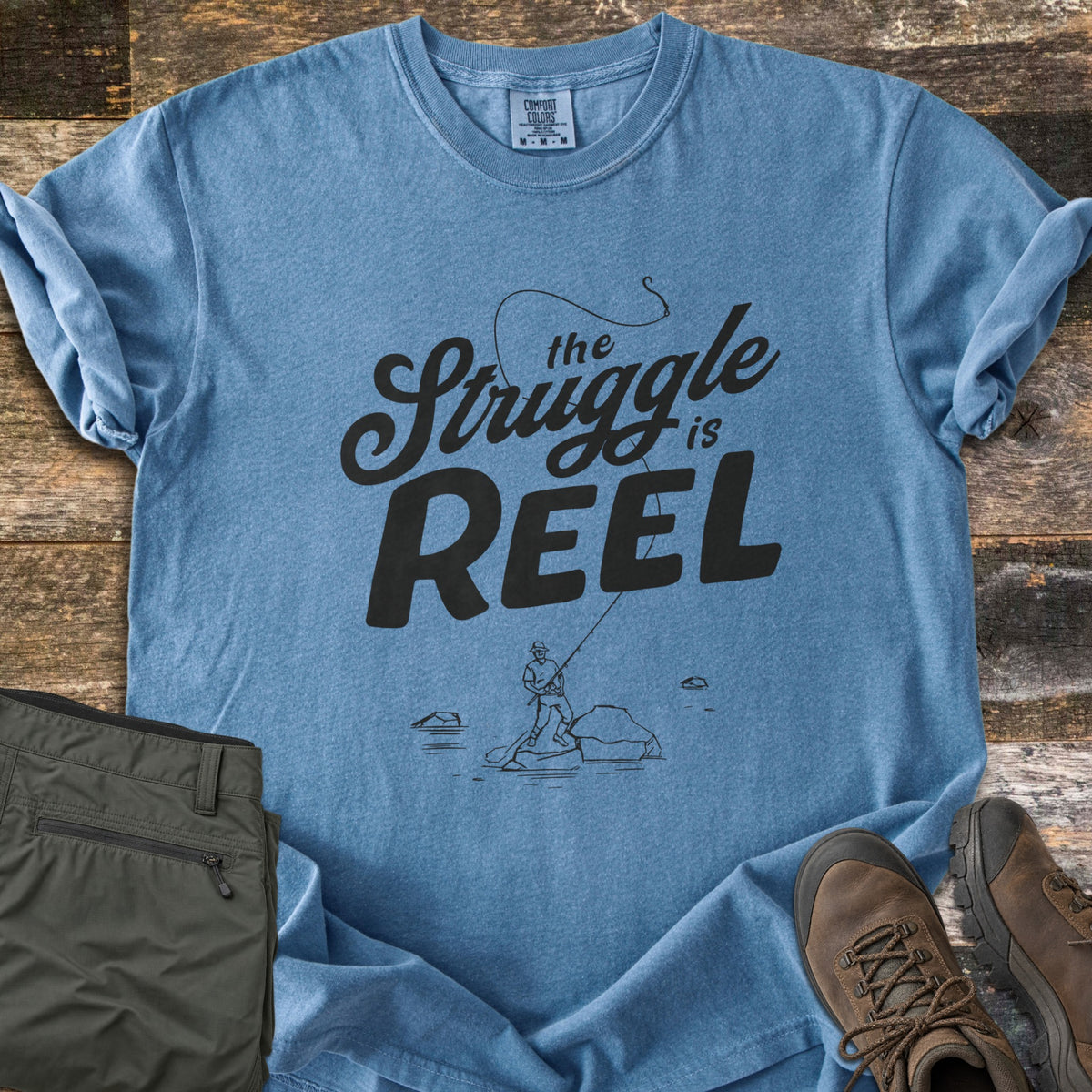 The Struggle Is Reel Comfort Colors T-shirt in Blue Jean, laid flat. Black text reads the Struggle is REEL under an arcing fishing line with a hook, above a sketch of an angler casting from a rock in water. Folded olive pants and brown hiking boots sit on wood planks.