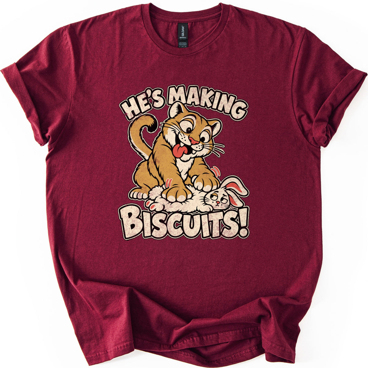 He's Making Biscuits Comfort Colors T-shirt in Maroon, laid flat. A grinning cartoon cougar with its tongue out kneads a dazed white rabbit on its back, with the words He's Making Biscuits! in distressed cream letters. The shirt lies on a white background with rolled sleeves.