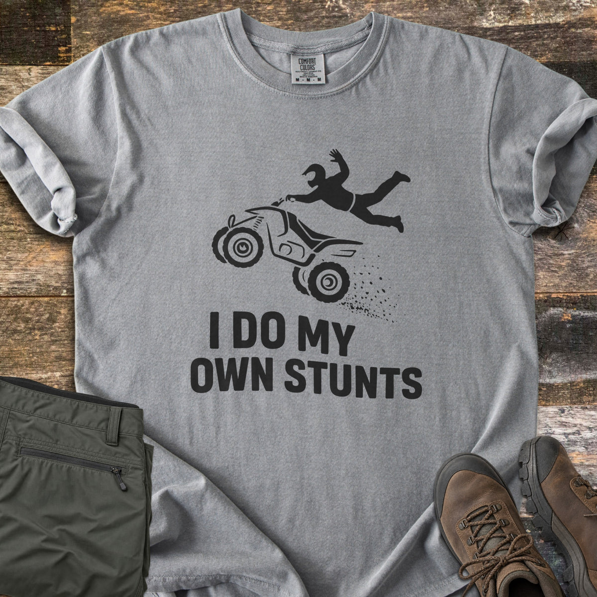 ATV Stunts Comfort Colors T-shirt in Grey, laid flat. A black silhouette of a helmeted rider flying off a wheelie-popping ATV kicking up dirt, above the words I Do My Own Stunts. Olive cargo pants and brown hiking boots sit beside it on weathered wood.